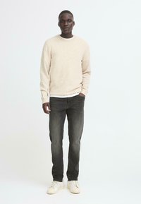 Bež pleteni pulover s okroglim izrezom, v kombinaciji s temno sivo jeans hlačami in svetlimi športnimi copati. Tkanina se zdijo mehka z teksturiranim zaključkom.