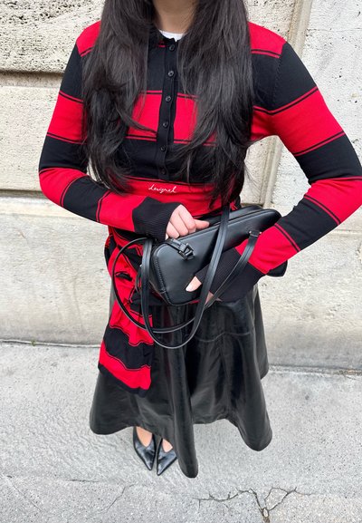 Mujer con una camiseta de manga larga de rayas rojas y negras y falda de cuero negra, sosteniendo un bolso negro frente a una pared de piedra.