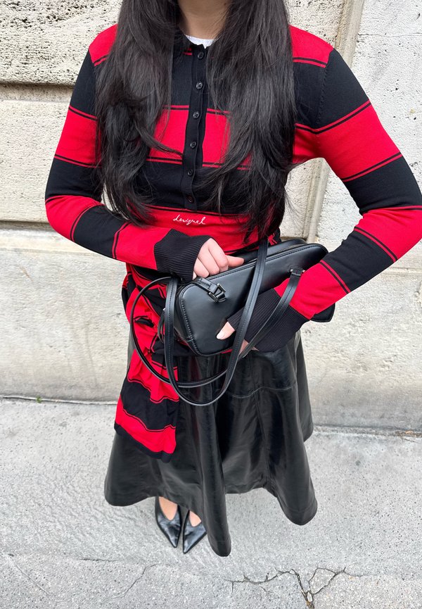 Kvinde iført en rød og sort stribet langærmet top og sort lædernederdel, holder en sort håndtaske foran en stenm væg.