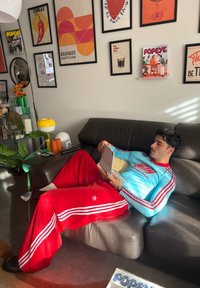 Άνδρας με μουστάκι και σκούρα μαλλιά κάθεται αναπαυτικά σε γκρι καναπέ χρησιμοποιώντας tablet, φορώντας ανοιχτό μπλε μπλουζάκι και κόκκινες φόρμες Adidas σε πολύχρωμο δωμάτιο.