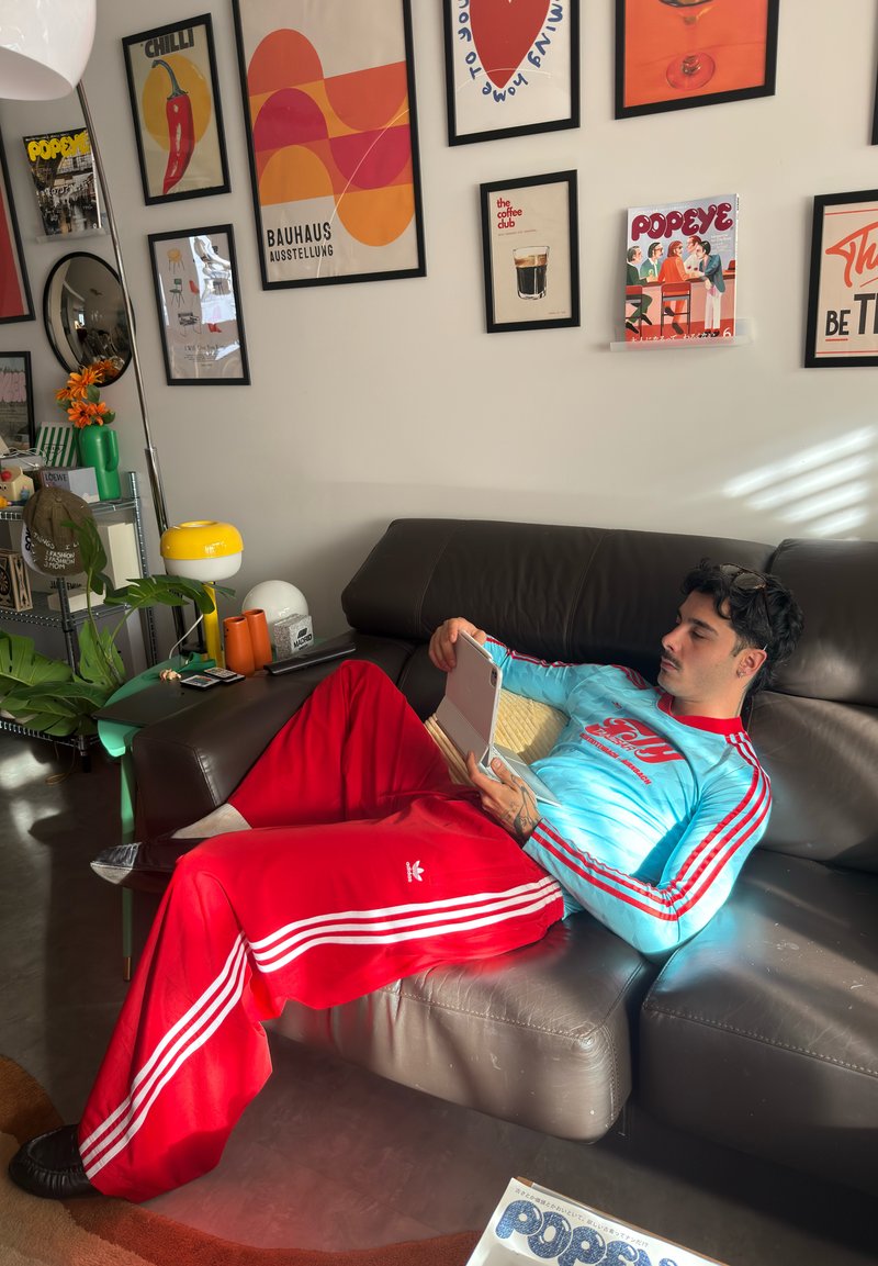 Homme avec moustache et cheveux foncés se détend sur un canapé gris en utilisant une tablette, vêtu d'un haut bleu clair et d'un pantalon de survêtement Adidas rouge dans une pièce colorée.