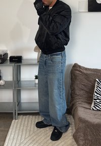 Must tikkimustriga pommitajajaq, helesinised laiade säärtega teksad ja mustad kingad. Kinnastel käed, seisab neutraalse tausta ees.