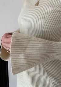 Χέρια ενωμένα πάνω σε κρεμ πλεκτό πουλόβερ με φαρδιά μανίκια καμπάνα και μικρό χρυσό μενταγιόν στο λαιμό.