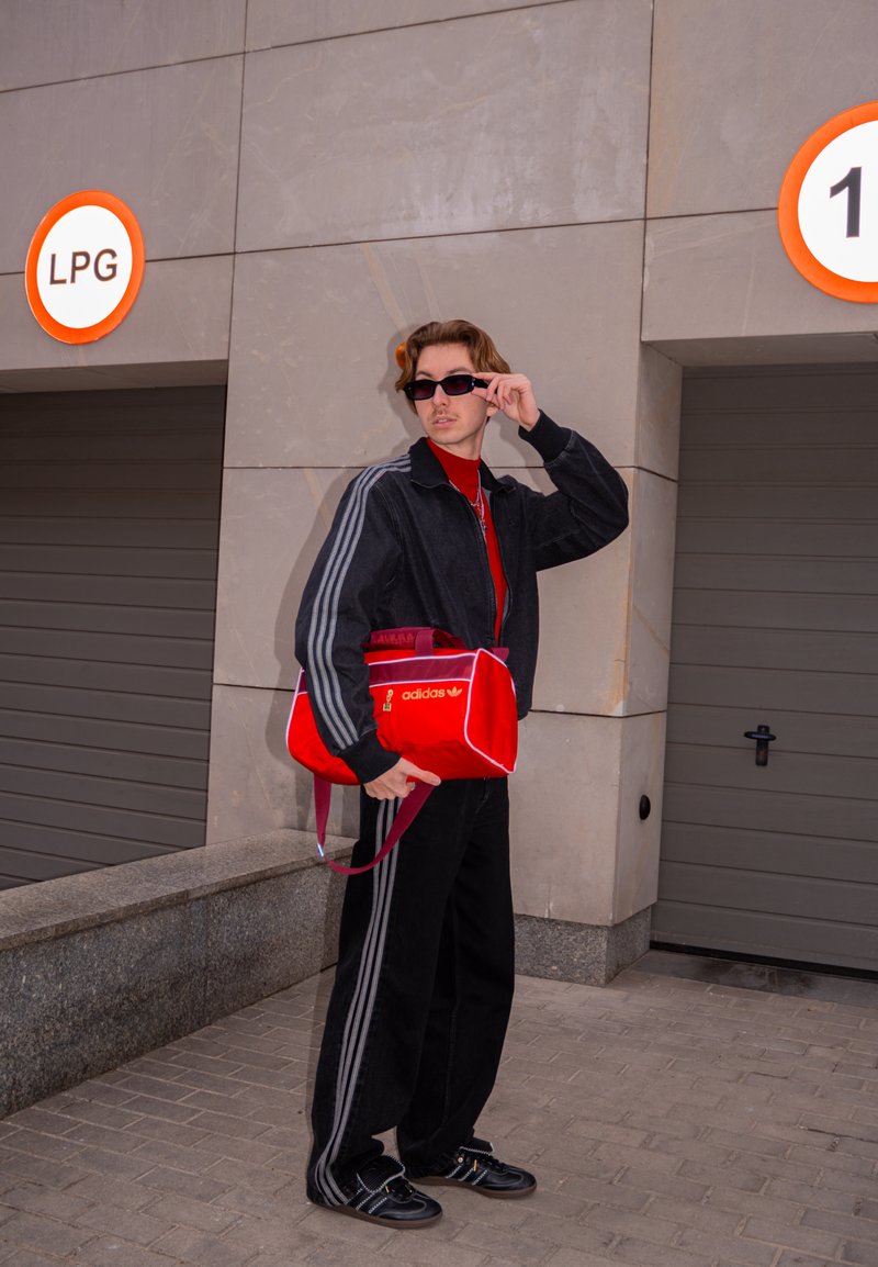 Fiatal férfi fekete Adidas melegítőben igazítja napszemüvegét, miközben egy piros Adidas táskát tart, szürke garázskapuk mellett áll, amelyeken "LPG" és "1" felirat szerepel.