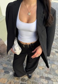 Zwarte blazer over een strak wit tanktopje, gecombineerd met wijde zwarte broek. Een textuur lichtgrijze clutch en een zilveren hangerketting met de letter 'N'.