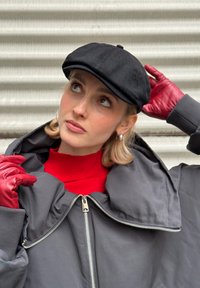 Cappello piatto nero, dolcevita rosso, giacca grigia oversize con collo alto e cerniera. Guanti in pelle rossa e orecchini a cerchio d'argento.