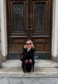 A wooden door with intricate metal accents serves as a backdrop. A person in a black coat and burgundy tights sits on stone steps.