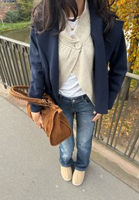 Marineblå blazer over en kremfarget teksturert topp med stor knapp, blå jeans, beige tøfler og en brun semsket skinnveske med flettet håndtak.