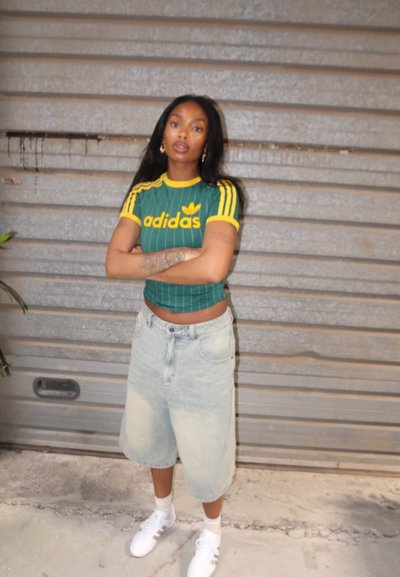 Top corto de Adidas a rayas verdes con acentos amarillos, pantalones cortos de mezclilla claros y sueltos, y zapatillas blancas. El cabello es largo y lo lleva suelto.