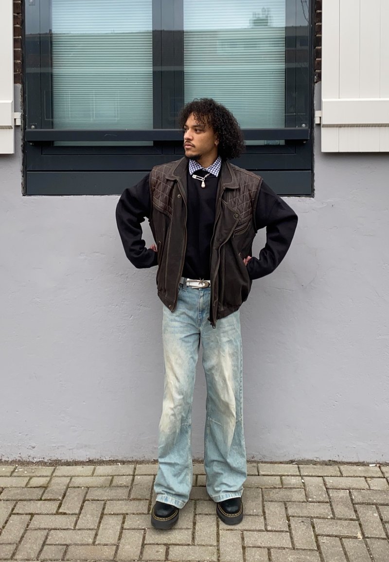 Barna steppelt mellény egy fekete pulóver felett, világoskék, bő szárú farmernadrággal és fekete platform csizmával párosítva. A háttérben egy szürke fal és ablakok láthatók.