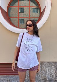 Biała koszulka z czarnym ręcznie pisanym tekstem "KOCHAM CIĘ NA ZAWSZE" oraz logo "Fiorucci", połączona z czerwoną i białą kratkowaną spódnicą. Brązowa torebka.
