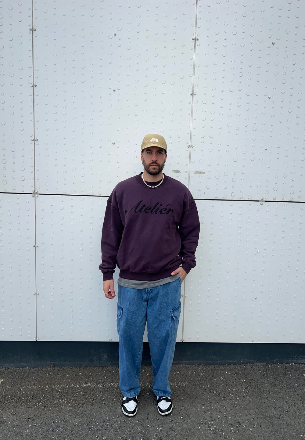 Sweatshirt violet avec le texte "Atelier", chemise à couches grise, pantalon cargo bleu ample, casquette beige, baskets noir et blanc, avec un collier en perles.