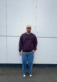 Sweatshirt violet avec le texte "Atelier", chemise à couches grise, pantalon cargo bleu ample, casquette beige, baskets noir et blanc, avec un collier en perles.