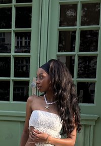 Top senza spalline in pizzo bianco, collana a forma di stella marina con perle, lunghi capelli ondulati e scuri, che sta vicino a porte in vetro con cornice verde.