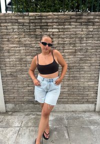 Musta lyhyt toppi, vaaleansiniset farkkushortsit repsottavilla helmalla, mustat varvassandaalit ja valkoiset kissansilmä-aurinkolasit teksturoidun tiiliseinän edessä.