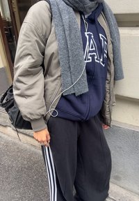 Pufová bunda v šedé, námořnická mikina s bílým logem, černé tepláky s bílými pruhy, šedý šátek a černá taška se stříbrnými detaily.