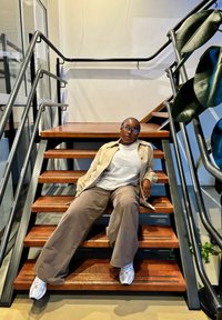 Ξύλινη σκάλα με σκούρα μεταλλική χειρολαβή; Ένα άτομο με μπεζ παλτό και χαλαρούς καφέ παντελόνες κάθεται στα σκαλοπάτια, με sneakers στα πόδια. Πράσινο φυτό κοντά.