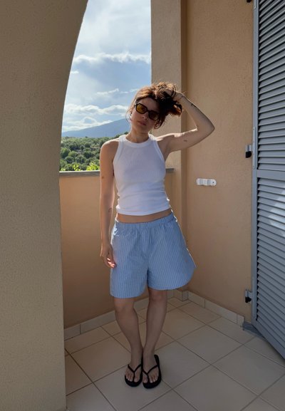 Camiseta sin mangas blanca de canalé, pantalones cortos a rayas azul claro con una cinturilla elástica, chanclas negras, de pie en un balcón con una pared texturizada.