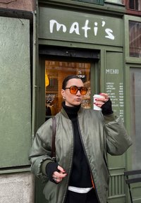 Personne portant des lunettes de soleil orange et une veste verte tenant un gobelet en papier devant l'entrée verte d'un café avec un menu visible.