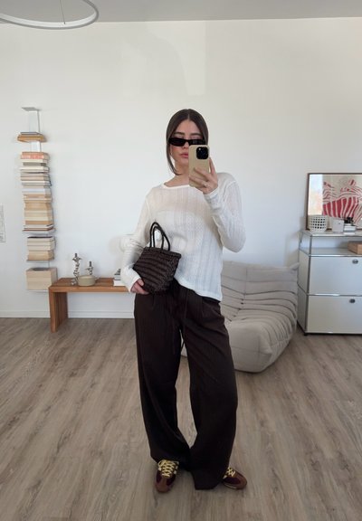 Suéter blanco de canalé, pantalones de pierna ancha a rayas marrones, bolso de mano negro trenzado y zapatillas marrones con cordones amarillos, en un interior minimalista.