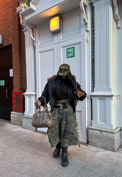 Persona con gorra y pantalones de camuflaje, chaqueta negra y botas, sosteniendo un bolso marrón claro, caminando junto a puertas blancas de salida de emergencia en la acera de la ciudad.