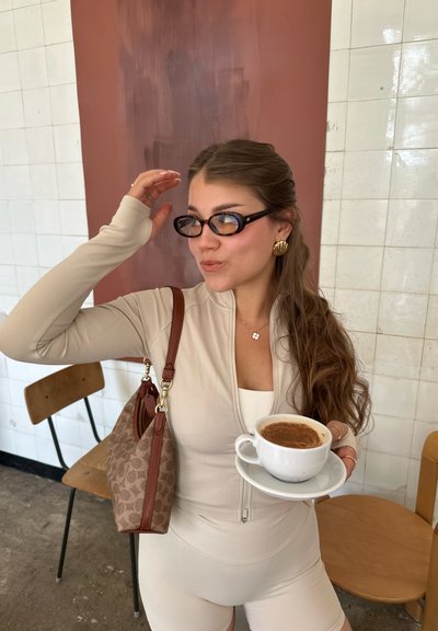 Bodysuit de canalé beige, gafas negras, pendientes de oro, sosteniendo una taza blanca con café en un plato, y un bolso marrón con un patrón.