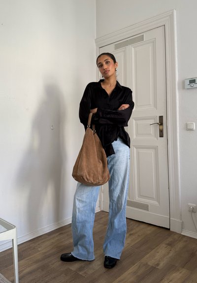 Mujer de pie en interiores con los brazos cruzados, vistiendo una blusa negra, jeans de pierna ancha de color azul claro, zapatos negros y sosteniendo un bolso grande de ante marrón.