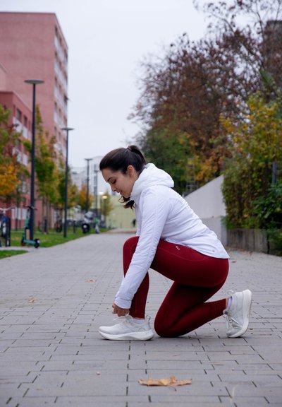 Chaqueta blanca con cierre, leggins rojos y zapatillas blancas. Vista a nivel del suelo de una persona arrodillada, atándose los cordones de los zapatos en un camino pavimentado bordeado de árboles.