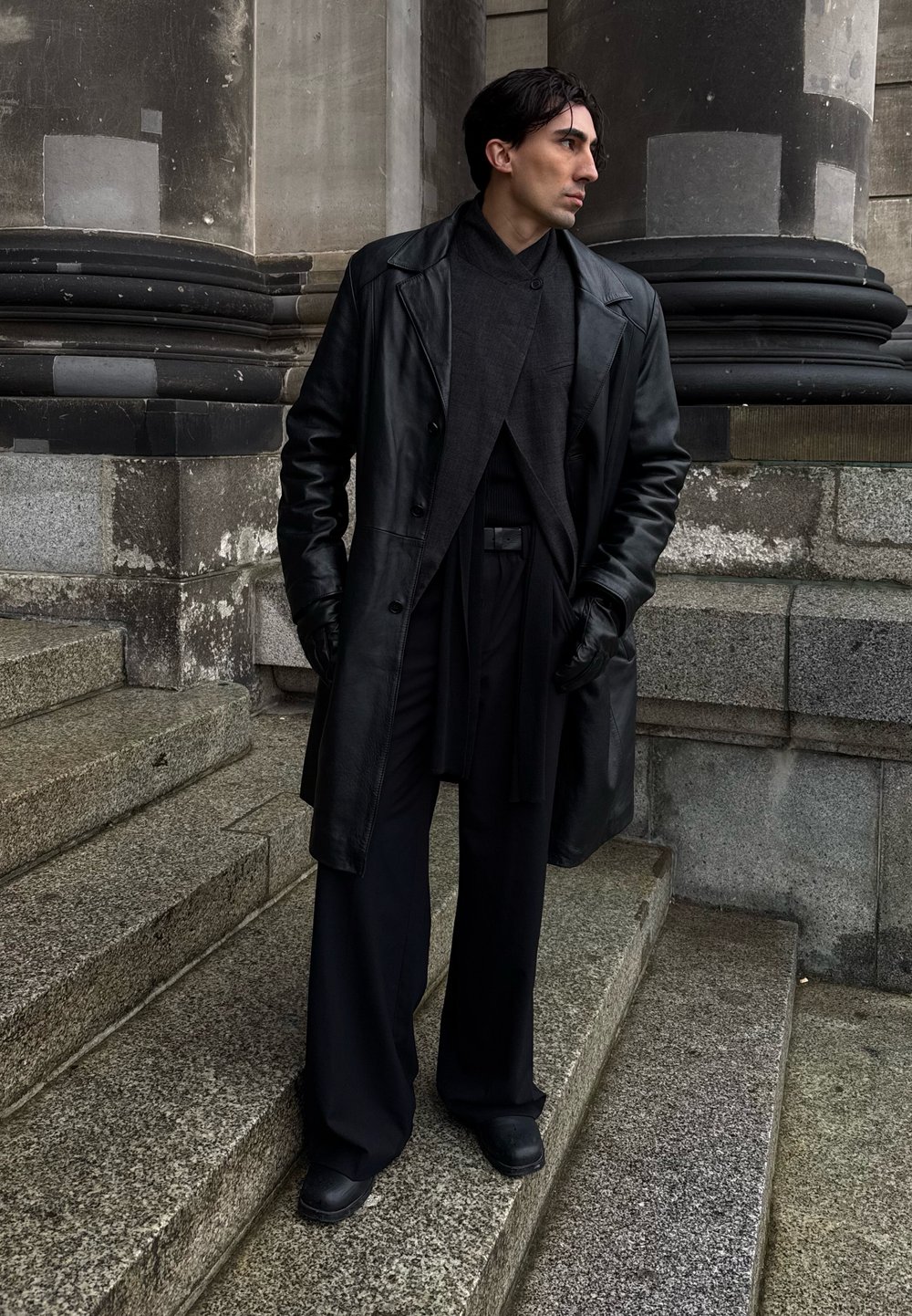 Mann med mørkt hår iført svart skinnfrakk, hansker og vide bukser står på steintrapper foran en bakgrunn av stein og søyler, og ser mot høyre.