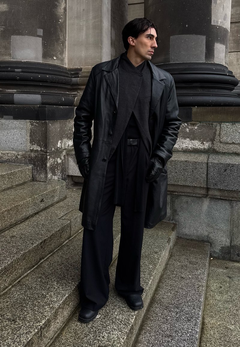 Uomo con capelli scuri che indossa un cappotto di pelle nero, guanti e pantaloni larghi, in piedi su gradini di pietra davanti a uno sfondo di pietra e colonne, guarda verso destra.