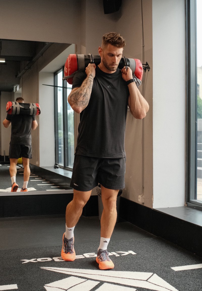Homme en tenue de sport noire portant un sac de sable lesté sur ses épaules à l'intérieur d'une salle de sport avec de grandes fenêtres et un mur miroir.
