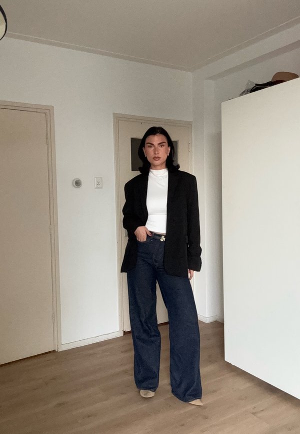 Mujer de pie en el interior que lleva una camiseta blanca, blazer negro, vaqueros anchos oscuros y zapatos beige sobre un suelo de madera cerca de puertas cerradas.