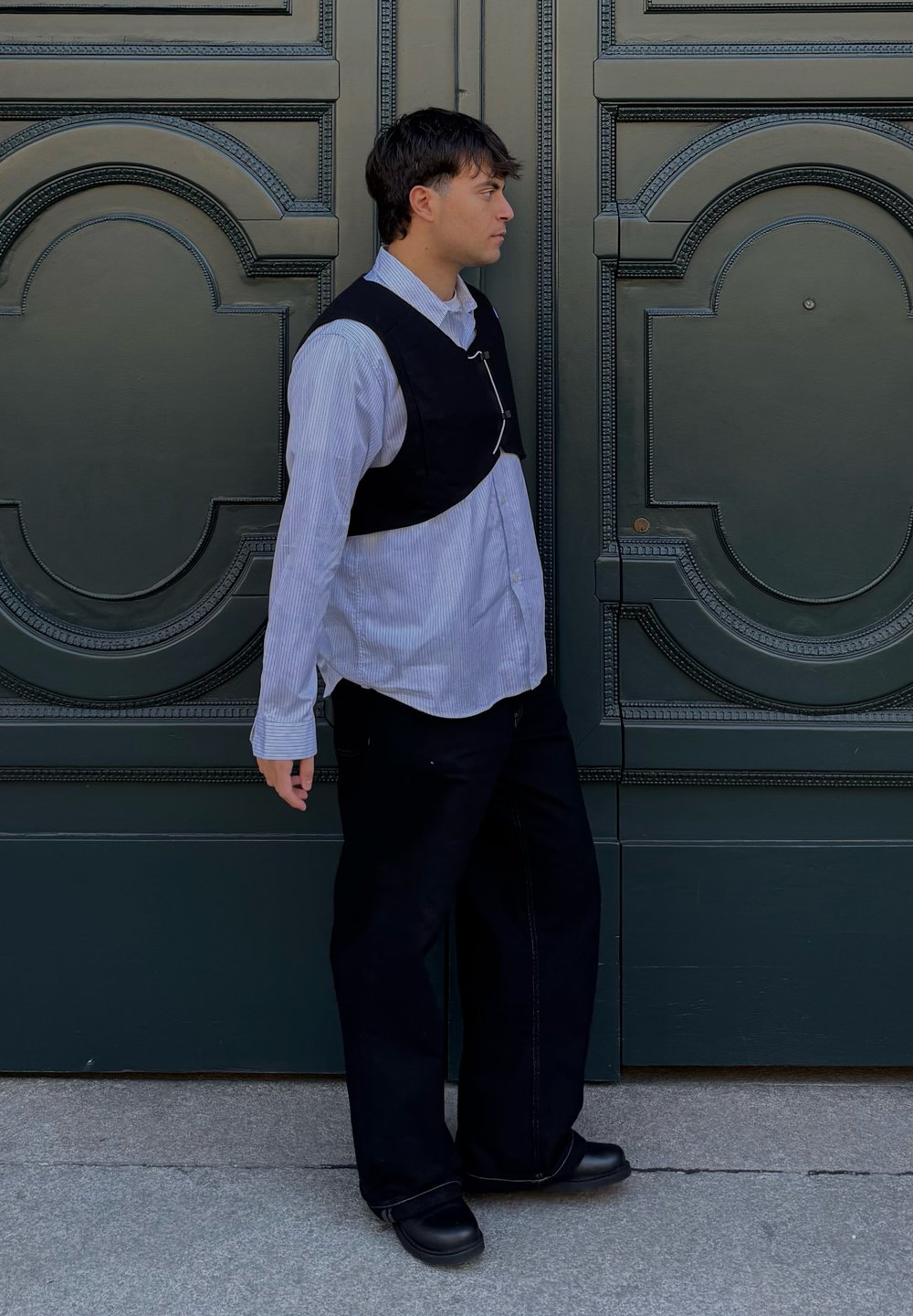 Jeune homme portant une chemise à rayures et un gilet noir, se tenant de profil contre des portes doubles vertes ornées de motifs circulaires.
