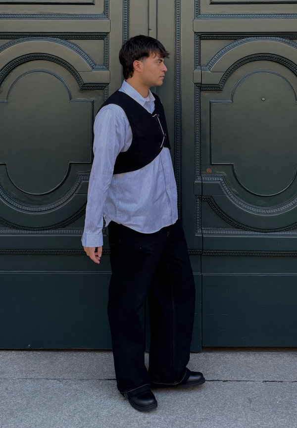 Mladić u prugastoj košulji i crnom prsluku stoji u stranu naslonjen na ukrasna zelena dvokrilna vrata s kružnim uzorcima.