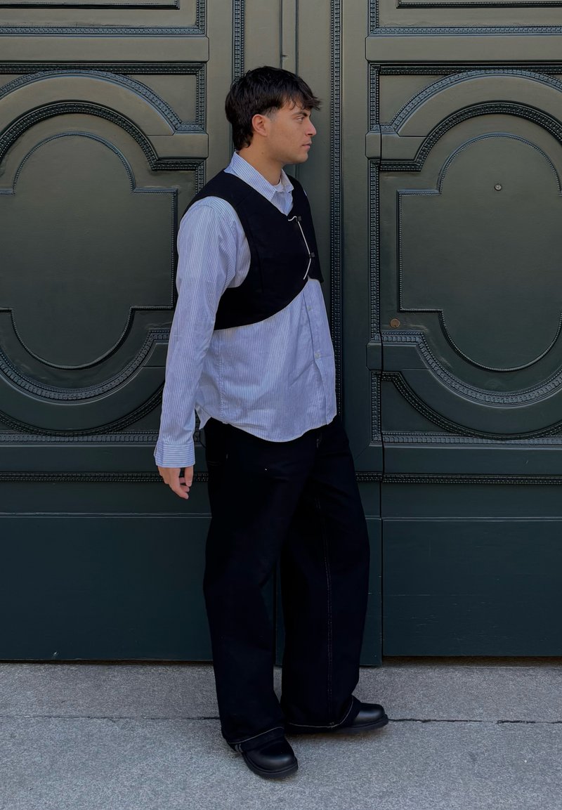 Jeune homme portant une chemise à rayures et un gilet noir, se tenant de profil contre des portes doubles vertes ornées de motifs circulaires.