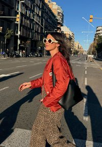 Raudona siuvinėta striukė, juoda odinė petneša, gyvūnų rašto kombinezonas, dideliai balti akiniai nuo saulės, ilgi rudieji plaukai, miesto gatvės fonas.