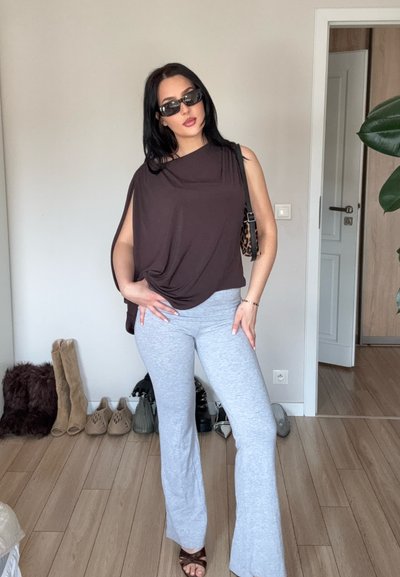 Mujer con camiseta negra sin mangas, pantalones grises, gafas de sol y sandalias posando en interior junto a una fila de zapatos variados contra una pared clara.