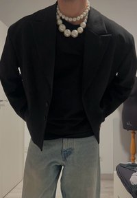 Zwarte blazer over een zwart shirt, gecombineerd met lichtblauwe spijkerjeans. Voorzien van grote parelkettingen als accessoires. Eenvoudig ontwerp.