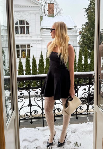 Mujer con vestido negro, medias de encaje y tacones, sosteniendo un bolso beige, de pie en un balcón cubierto de nieve con barandilla ornamentada, árboles nevados y un edificio blanco al fondo.