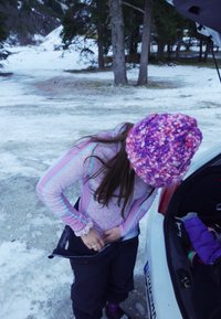 Kvinne iført lys lilla langermet skjorte og tykk, strikket rosa-lilla lue, justerer svarte skibukser nær en bil i snødekt terreng.