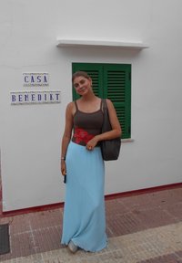 Žena stojí vedľa steny s modro-bielymi znamení. Má na sebe priliehavé hnedé tričko s červenou kvetinou, svetlomodrú maxi sukňu a drží tašku.