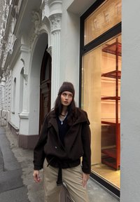 Manteau en laine marron avec ceinture à nouer, pull noir en dessous, et pantalon beige. En arrière-plan, une vitrine avec des étagères rouges.