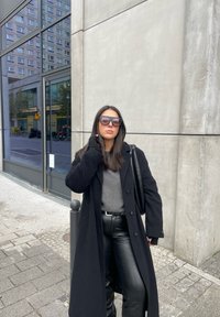 Μαύρο μάλλινο παλτό πάνω από ένα γκρι πουλόβερ, συνδυασμένο με μαύρο δερμάτινο παντελόνι. Μεγάλα μαύρα γυαλιά ηλίου, στέκεται μπροστά από έναν τσιμεντένιο τοίχο, με φόντο την πόλη.