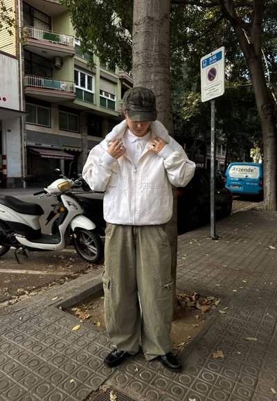 Chaqueta bomber blanca con cuello de piel, pantalones cargo verdes y gorra de camuflaje, de pie junto a un scooter en una calle de la ciudad.