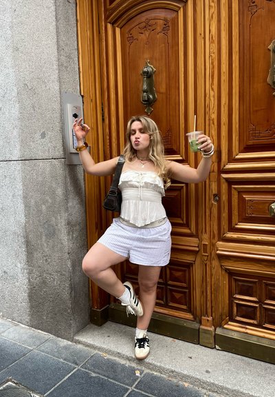 Top peplum blanco con volantes, pantalones cortos a rayas claros y zapatillas negras. De pie al lado de una puerta de madera con detalles ornamentados. Sosteniendo una bebida verde.
