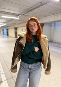 Veste en fausse peau lainée marron avec doublure en fausse fourrure claire, pull vert et jean délavé clair ; l'environnement est un garage en béton.