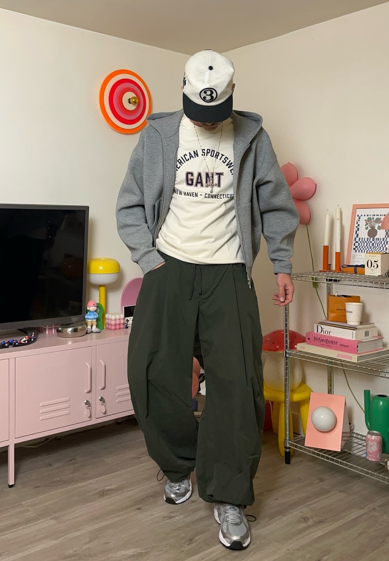 Felpa con cappuccio grigia con cerniera, t-shirt bianca con testo rosso, pantaloni verdi oliva oversize e scarpe da ginnastica grigie. Accessori includono un cappello nero e bianco.