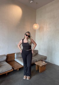 Sort tætsiddende tanktop og bukser, begge i et strækbart materiale. Bløde grå sofaer og et træbord er i baggrunden.