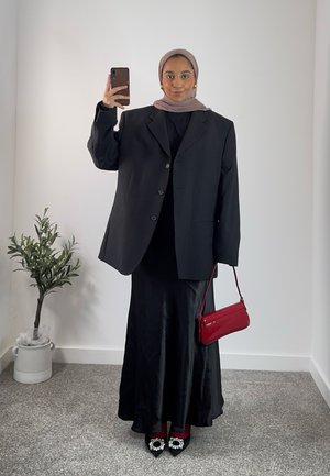 Blazer negra de gran tamaño, falda larga de satén negro, bolso rojo de charol, tacones negros con detalles de perlas. Fondo liso con una pequeña planta.