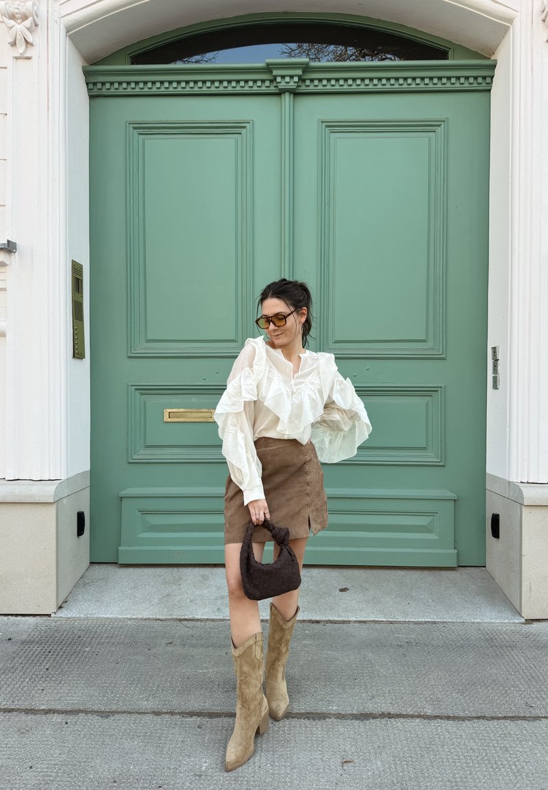 Femme portant une jupe en daim marron, un chemisier blanc à volants, des bottes de cow-boy couleur sable et des lunettes de soleil, tenant un sac à main marron foncé, debout devant des portes doubles vertes.